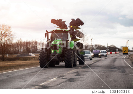 Large green agricultural tractor with specialized planting equipment travels on asphalt road, followed by several passenger cars, rural background. Copy space for text 138165318