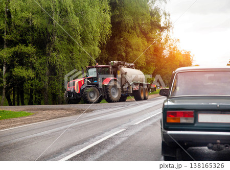 Red tractor pulls liquid tank trailer, country road, green trees line route, sun glow, view from passenger vehicle rear, clear day. Red tractor pulls liquid tank trailer, country road, green trees line route, sun glow, view from passenger vehicle rear, clear day. 138165326