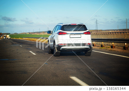 White SUV driving on asphalt highway road under clear blue sky with power lines and green fields in background during bright sunny day journey travel transport concept White SUV driving on asphalt highway road under clear blue sky with power lines and green fields in background during bright sunny day journey travel transport concept 138165344