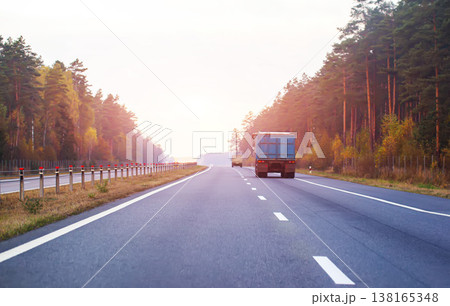 Highway with trucks driving through forest during day, guardrail roadside, open road travel, long distance transportation journey with bright sky 138165348
