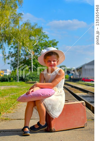 A sweet little girl in a white dress and hat is waiting for her holidays. The little girl is waiting for the holiday train to arrive. The concept of a carefree childhood, summer holidays, and family. A sweet little girl in a white dress and hat is waiting for her holidays. The little girl is waiting for the holiday train to arrive. The concept of a carefree childhood, summer holidays, and family. 138165468