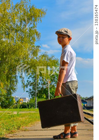 A cute little boy on a train station platform. A sad little boy in a flat cap holds an old suitcase. The concept of travel, vacation, and a carefree childhood. Retro and vintage vibe. A cute little boy on a train station platform. A sad little boy in a flat cap holds an old suitcase. The concept of travel, vacation, and a carefree childhood. Retro and vintage vibe. 138165474
