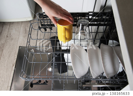 Human hand places yellow ceramic mug into upper rack of modern kitchen dishwasher for cleaning, demonstrating efficiency of household appliance Human hand places yellow ceramic mug into upper rack of modern kitchen dishwasher for cleaning, demonstrating efficiency of household appliance 138165633