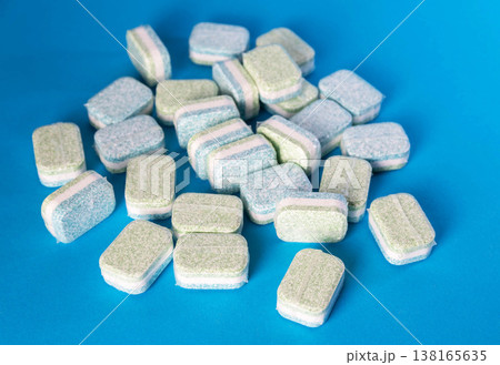 Pile of multi-layered dishwasher tablets individually wrapped in plastic on a vibrant blue background. Rectangular cleaning capsules for kitchen hygiene. 138165635