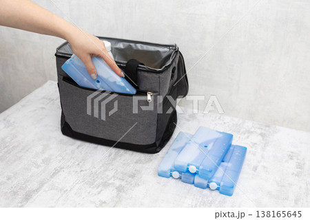 Woman hand placing blue rectangular ice pack inside open grey fabric cooler bag. Multiple cold generators next to bag on light textured surface, preparation for travel or picnic. Woman hand placing blue rectangular ice pack inside open grey fabric cooler bag. Multiple cold generators next to bag on light textured surface, preparation for travel or picnic. 138165645