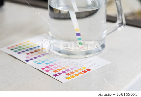 Water quality test strip measurement of parameters in glass pitcher with clear liquid, comparing results with color chart for accurate analysis of water composition. 138165655