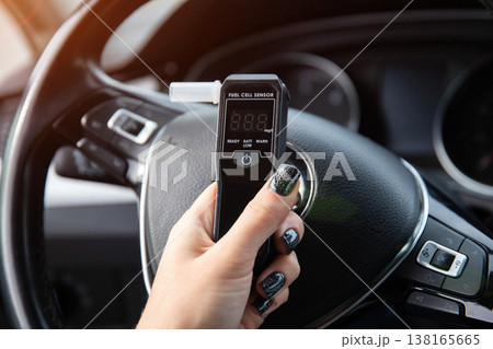 Woman's Hand Holding Breathalyzer Over Steering Wheel. Female Driver Checks Sobriety Before Trip. 138165665