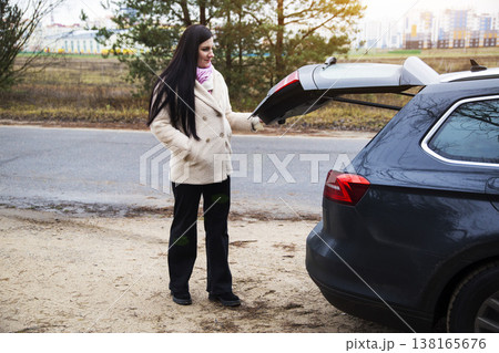 Woman standing next to car with open trunk on roadside, preparing for travel or unpacking, city buildings and pine trees in background, daytime 138165676