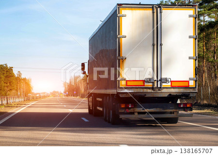 Rear view of white semi-trailer truck driving rural highway. Cargo transport vehicle travels open road, clear sky, long shadows alongside asphalt pavement. 138165707