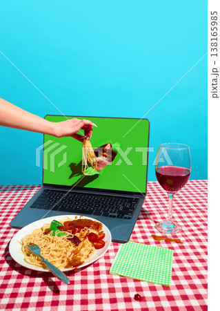 Hand feeding spaghetti into mouth emerging from laptop screen on table with wine. Concept of food delivery platform, digital consumption, online service advertising. Hand feeding spaghetti into mouth emerging from laptop screen on table with wine. Concept of food delivery platform, digital consumption, online service advertising. 138165985