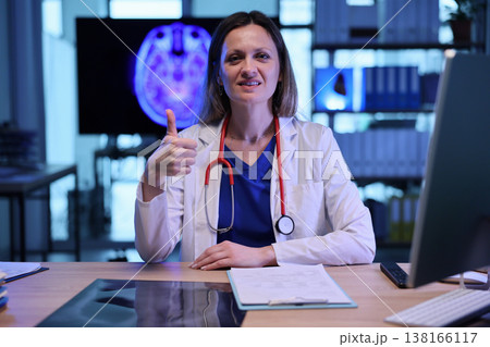 Confident female doctor at her desk in a modern medical office shares positive message during a patient consultation 138166117