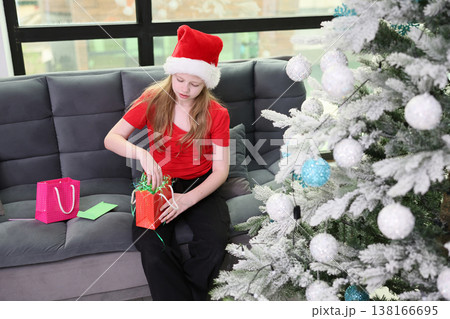 Excitement fills the air as a child in a festive hat unwraps a gift beside a sparkling Christmas tree in a cozy living room Excitement fills the air as a child in a festive hat unwraps a gift beside a sparkling Christmas tree in a cozy living room 138166695