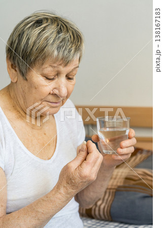 Senior woman carefully holding a pill and a glass of water, focusing on daily medication for health and wellness. Health care, prevention of age-related diseases. Senior woman carefully holding a pill and a glass of water, focusing on daily medication for health and wellness. Health care, prevention of age-related diseases. 138167183