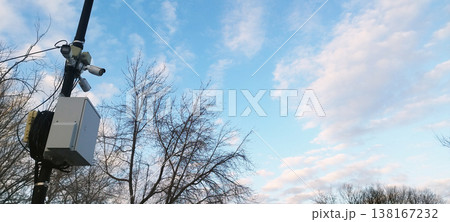 A high pole holds multiple white surveillance cameras and a grey electrical box. Bare trees are set against a blue sky with soft white clouds, symbolizing constant observation in a public space 138167232