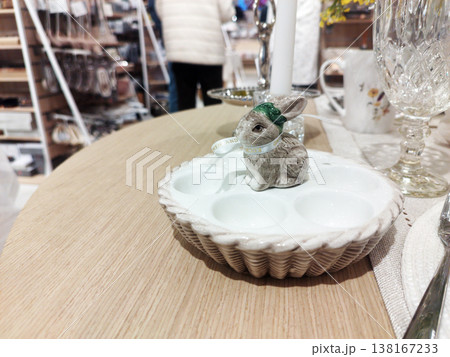 Small, realistic bunny figurine with green headband, ribbon sits in the center of ceramic egg tray. The basket-weave patterned dish is displayed on wooden table within bright home goods store. Easter 138167233