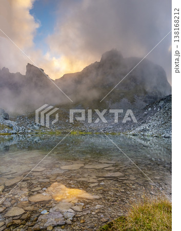 Mist rises over a serene Wildsee lake in the Pizol mountains at sunrise, casting a mystical atmosphere. The rocky terrain and clear waters reflect the early morning light golden hour. Mist rises over a serene Wildsee lake in the Pizol mountains at sunrise, casting a mystical atmosphere. The rocky terrain and clear waters reflect the early morning light golden hour. 138168212