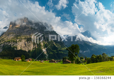 Majestic mountains rise in the background as quaint wooden cabins dot green landscape. A winding path guides a visitor through this serene Swiss alps. Grindelwald Jungfrau Bernese alps Switzerland. Majestic mountains rise in the background as quaint wooden cabins dot green landscape. A winding path guides a visitor through this serene Swiss alps. Grindelwald Jungfrau Bernese alps Switzerland. 138168244