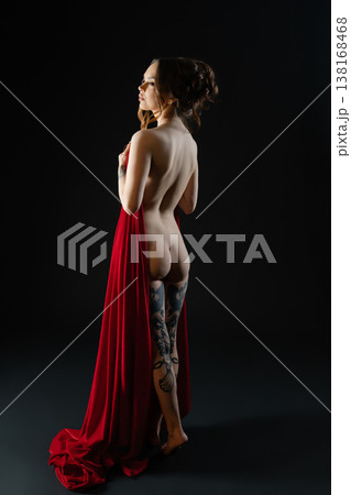 Elegant tattooed woman seen from behind, holding flowing red fabric against a dark studio background. Elegant tattooed woman seen from behind, holding flowing red fabric against a dark studio background. 138168468