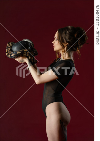 Profile of an elegant tattooed woman in a sheer bodysuit holding a python snake wrapped around a black sphere. 138168476