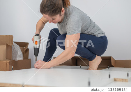 A Caucasian woman uses a screwdriver to tighten a screw while assembling a cabinet. A Caucasian woman uses a screwdriver to tighten a screw while assembling a cabinet. 138169184