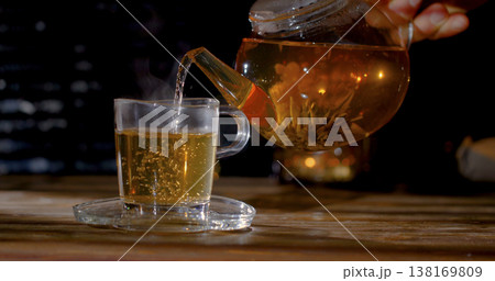 Warm tea is poured from a teapot into a clear glass cup at a cozy setting during evening hours 138169809