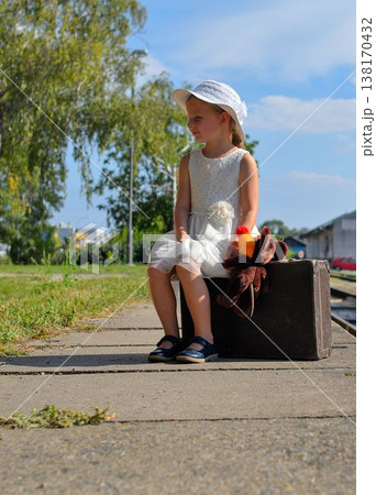 A little girl on a train platform. A cute girl at a train station. A little girl holds a stuffed toy and sits on an old suitcase. The concept of travel, holidays, and a carefree childhood. Retro and A little girl on a train platform. A cute girl at a train station. A little girl holds a stuffed toy and sits on an old suitcase. The concept of travel, holidays, and a carefree childhood. Retro and 138170432