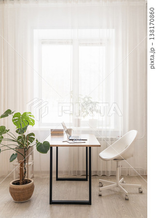Modern home office workspace featuring laptop on wooden desk with white mug and notebook, surrounded by green plants bookshelves 138170860