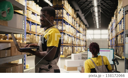 Black male staff verifying inventory pallets in e-commerce logistics center. Accurate order processing on laptop and packaging for quick shipping in large scale warehouse setting. Camera A. 138172066