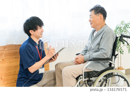 車椅子に乗るシニア男性を問診する介護職の男性スタッフ 138176571