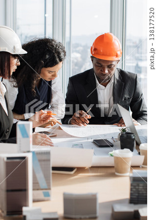 A diverse team of architects wearing hard hats collaborates around a table, reviewing construction plans and building models 138177520