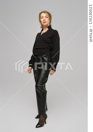 Middle-aged woman poses in black outfit against a plain gray backdrop during a fashion shoot 138180023