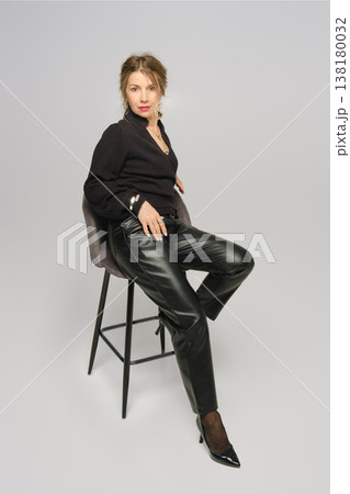 Model poses on a stool wearing black clothing in a studio setting during the day 138180032