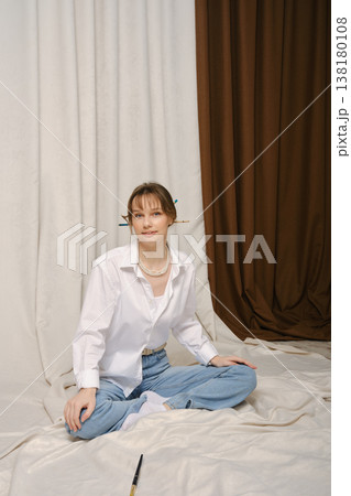 Young woman sitting on white fabric, wearing casual clothes and a necklace in a studio setting 138180108