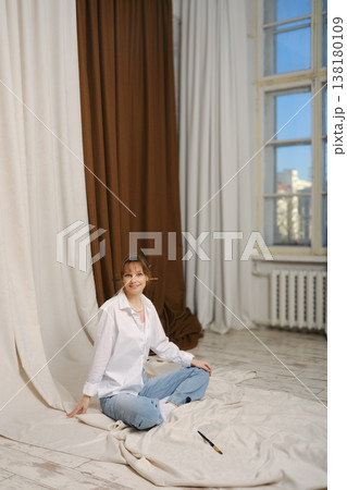 Smiling girl sitting on the floor in a studio surrounded by fabric backdrops 138180109