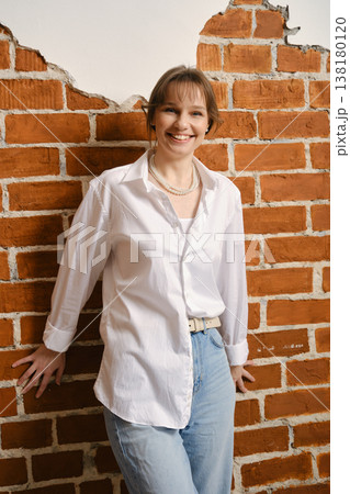 Smiling person leans against a brick wall wearing a white shirt and light jeans in a casual setting 138180120