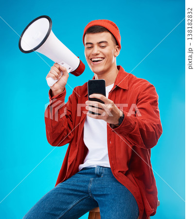 Phone, megaphone and young man in a studio reading announcement or speech for a rally. Happy, cellphone and male activist or protest with bullhorn for loud communication isolated by blue background 138182832