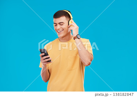Phone headphones and young man in a studio listening to music, radio or playlist for entertainment. Smile, technology and male person streaming a song, podcast or album isolated by blue background. 138182847