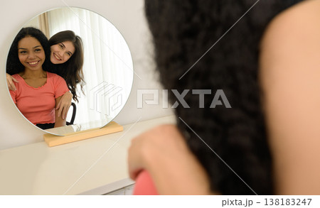 Two smiling couple women enjoying time together in front of mirror. Asian female hugging African woman from behind. Two smiling couple women enjoying time together in front of mirror. Asian female hugging African woman from behind. 138183247