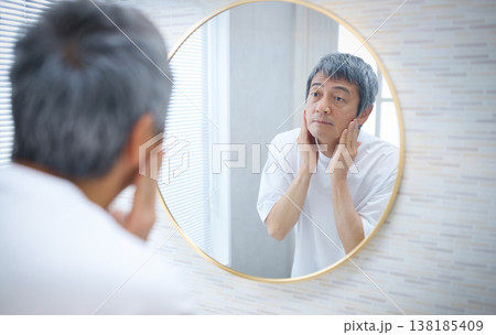 鏡を見ながら顔を手で触る60代の男性　 138185409