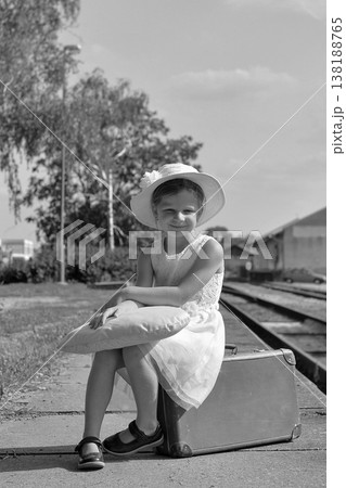 Young girl in a white dress and hat sits on a suitcase at a train station platform Young girl in a white dress and hat sits on a suitcase at a train station platform 138188765