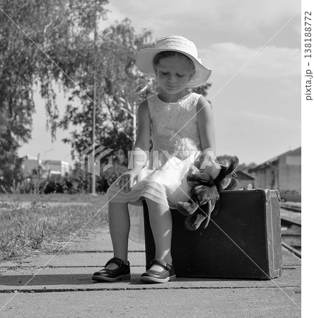 Young girl in a hat and dress sits on a suitcase with a toy, waiting patiently Young girl in a hat and dress sits on a suitcase with a toy, waiting patiently 138188772