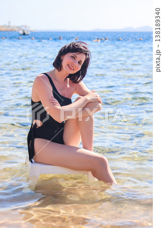 Elegant woman in a black swimsuit sitting on a plastic chair, enjoying the serene sea view, capturing the essence of summer relaxation and beach vibes Elegant woman in a black swimsuit sitting on a plastic chair, enjoying the serene sea view, capturing the essence of summer relaxation and beach vibes 138189340