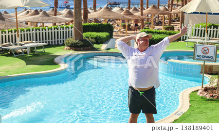 Man standing by the pool with arms behind his head, enjoying the warm sun, embodying relaxation and tranquility in a serene outdoor setting Man standing by the pool with arms behind his head, enjoying the warm sun, embodying relaxation and tranquility in a serene outdoor setting 138189401