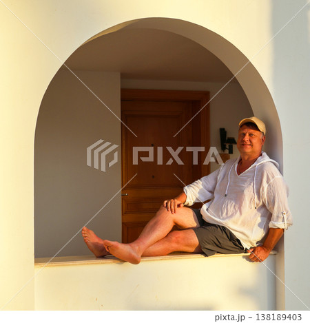 Adult man in a white shirt and cap sitting on a wide windowsill, enjoying the beautiful sunset, reflecting on life and tranquility in the golden hour Adult man in a white shirt and cap sitting on a wide windowsill, enjoying the beautiful sunset, reflecting on life and tranquility in the golden hour 138189403