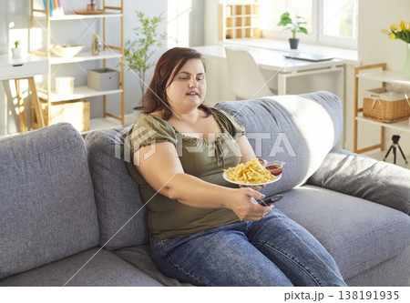Fat overweight woman sitting on sofa at home watching tv and eating tasty french fries. 138191935
