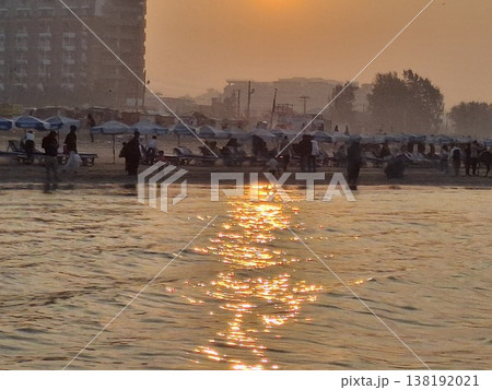 Golden Sunset Over Crowded Beach With Sun Reflection on Water and City Silhouette Golden Sunset Over Crowded Beach With Sun Reflection on Water and City Silhouette 138192021
