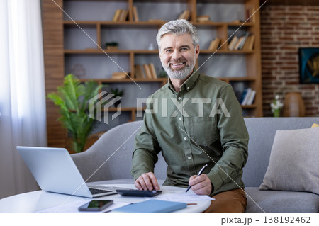 Smiling mature man on a sofa in a cozy living room, using laptop and calculator to review bills, budget, and retirement planning while working on paperwork at home 138192462