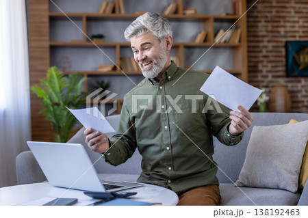 Senior man smiling excitedly while reviewing paperwork and using a laptop at a home office setup, celebrating financial success and managing bills online Senior man smiling excitedly while reviewing paperwork and using a laptop at a home office setup, celebrating financial success and managing bills online 138192463