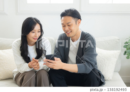 ソファーに座ってスマホを見る夫婦　 138192493