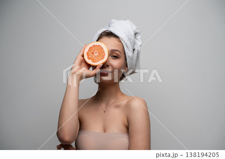 Young woman enjoying natural beauty and fresh spa wellness after a shower, holding an organic grapefruit half over her eye, promoting healthy skincare 138194205